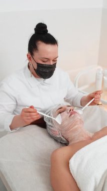 Enzim terapisi. Güzellik uzmanı, genç bir kadının yüzüne fırçayla nemlendirici krem maske takıyor. Kaplıcadaki kız cilt bakımı prosedürü. Yüz kozmetolojisi tedavisi. Dikey video görüntüsü