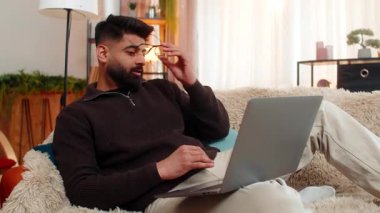 Evde oturan Hintli adam, dizüstü bilgisayar kullanırken yorgun bir ifadeyle gözlerini ovuşturuyor, uzun ekran görüntüsünden yorulmuş. Kanepedeki Arap adam rahatsızlık ve göz yorgunluğu gösteriyor. Dijital aşırı stresle boğuşuyor.