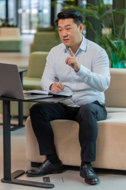 Genç Çinli iş adamı, otel lobisindeki internet video dersi veya ofis toplantısına katılırken not alıyor ya da not alıyor. Web kamerasına bakan gömlek destek servislerinde kendine güvenen bir yönetici