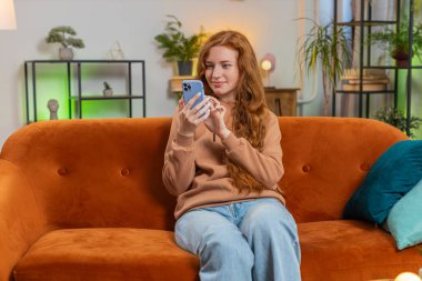 Evdeki oturma odasında gülümseyen cep telefonu kullanan kızıl saçlı mutlu bir genç kadın. Akıllı telefon sosyal medya uygulamalarında internetten film izleyen kız mesajları paylaşıyor.