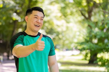 Gibi. Portre atletizm formundaki gülümseyen koşucu eğitimi maraton başarısından dolayı mutlu görünüyor. Parkta egzersiz kardiyolojisi. Japon genç sporcu bulanık ağaçlara karşı baş parmağını kaldırıyor