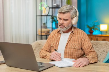 Beyaz, olgun bir adam evde oturmuş kulaklıklı bir dizüstü bilgisayardan podcast ya da online yayın dinliyor. Adam not defterine not tutuyor, içerikle ilgileniyor, eğitimi öğreniyor.