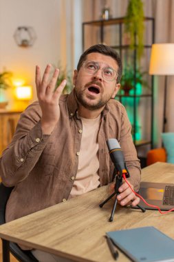 Genç, beyaz, gözlüklü bir blog yazarı mikrofona şarkı söylerken dizüstü bilgisayar kullanıyor ve eliyle ritmik hareketler yapıyor. Oturma odasında oturuyor..