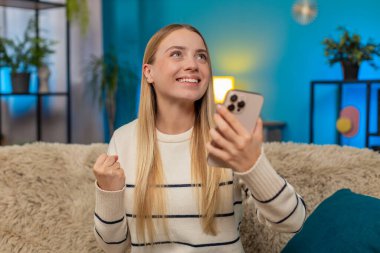 Beyaz bir kadın evdeki kanepede oturmuş online oyunu kutluyor ve zaferi kazanıyor. Heyecanlı kız akıllı telefonu tutuyor, sevinçle el kaldırıyor, gülümsüyor, mutluluğu, heyecanı ve başarıyı içeride hissediyor.