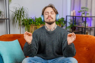 Meditasyon, dinlenme, mola. Müzik dinleyen beyaz adam derin derin nefes alıyor, gözleri kapalı meditasyon yapıyor konsantre düşüncelerle, huzurlu zihinle. Genç adam evdeki kanepede oturuyor.