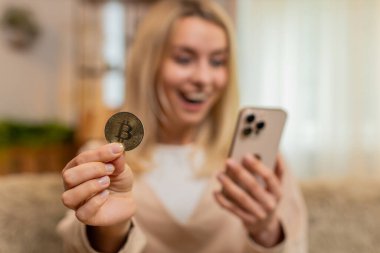 Genç yetişkin bir kadın evdeki kanepeden dizüstü bilgisayardan kripto grafiğini kontrol ediyor ve bitcoin dalgalanmasını kutlamak için elini kaldırıyor. Sarışın kız gururla gülümsüyor, yatırım kazancının ve piyasa başarısının zaferinin tadını çıkarıyor..