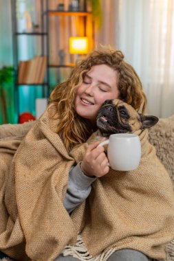 Beyaz saçlı genç bir kadın elinde sıcak ve sakin bir bardakla battaniye altında çay içiyor. Pug dog aynı battaniyenin altında huzur ve huzur dolu bir aile anını paylaşıyor..
