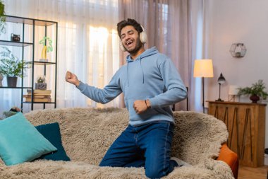 Kablosuz kulaklıklı genç adam kanepede dans ediyor, enerjik disko müziği dinliyor, hayali gitar çalıyor. İnsanlar hafta sonu eğlence aktivitelerinin keyfini çıkarıyorlar. İspanyol adam hava gitarıyla eğleniyor..