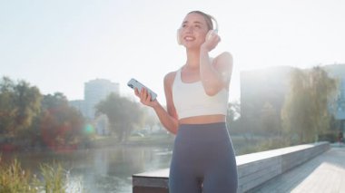 Atletik fitness sporu koşucusu kız antrenmanı, akıllı telefon tutmak kulaklıkla müzik dinlemek, dans etmek, eğlenmek. Parkta egzersiz kardiyolojisi. Danstan hoşlanan genç bir bayan koşucu. Aktif sporcu kadın