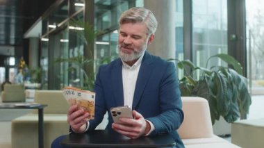 Başarılı, mutlu, olgun bir iş adamı akıllı telefon tutuyor ve ofis lobisinde oturmuş euro banknotlarını sayıyor. İnternette piyango kazanan beyaz sakallı adam. Bütçe hesaplanıyor.