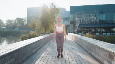 Güneşli bir günde şehir parkında köprüde atlayan beyaz tenli genç bir kadın. Dışarıda egzersiz yapan aktif formda bir bayan koşucu. Sağlıklı bir yaşam tarzı ve açık hava aktivitesi. Zindelik kavramı