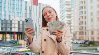 Zengin, zengin, beyaz, beyaz bir iş kadını akıllı telefon kullanıyor ve şehir merkezindeki bir sokakta elinde para dolu banknotlar tutuyor. Başarılı iş kariyeri, piyango kazananı, büyük gelir, zenginlik