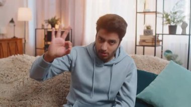 Neşeli Hintli adam kameraya olumlu bakıyor, iyi bir jest yapıyor, işaret gibi olumlu. Modern dairede koltukta oturan Arap Hintli adam, kendine güvenerek gülümsüyor, iyi bir şeyi onaylıyor..