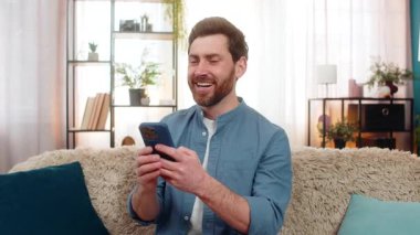 Genç adam akıllı telefondan komik sosyal medya yorumlarını okurken gülüyor. Kanepede oturan mutlu beyaz adam, elinde cep telefonu, çevrimiçi içerik etkileşimleri videolarını izlemekten zevk alıyor.