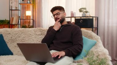 Evdeki Hintli adam dizüstü bilgisayarda odaklanmış bir ifadeyle yaklaşan online sınava tüm dikkatle hazırlanıyor. Kanepedeki Arap adam başarı için çalışmalara ve materyalleri ezberlemeye odaklanıyor.