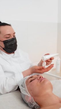 Güzellik uzmanı, genç yetişkin kadın yüzüne nemlendirici tıbbi kimyasallar soyarak krem fırçalama maskesi uyguluyor. Dişi cilt bakımı prosedürü. Yüz kozmetolojisi cilt yenilenmesi. Dikey