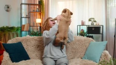 Genç Kafkasyalı kızıl saçlı kadın pug dog 'u heyecan ve zafer dolu bir gülümsemeyle evdeki kanepeye kaldırıyor. Evcil hayvan dalgaları, her ikisi de kapalı alanlarda paylaşılan enerjik kutlama anlarında neşeyi ifade ederken neşeyle pençelerini sallar..