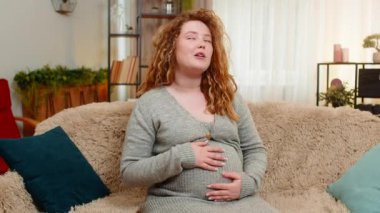 Hamile genç bir kadın kanepesinde oturup karnına hafifçe dokunuyor ve sevgi dolu bir ifadeyle yumuşakça konuşarak gelecekteki bebekle erken bir bağ kuruyor. Kız duygusal anları kucaklıyor, sıcak bakımı hissediyor.