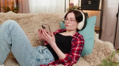 Genç kadın evdeki kanepede kablosuz kulaklık takıyor, derin bir ilgiyle sesli kitap dinlerken başını sallıyor. Kız hikayeye dikkatle ve odaklanarak tepki veriyor, ilgi çekici hikaye deneyiminden zevk alıyor..