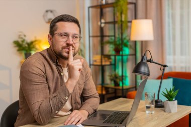 Sessiz olun lütfen. Kafkasyalı genç işadamı işaret parmağını dudaklarına bastırıyor. Ofis masasında sessiz jest işareti yapıyor. Oturma odasında dizüstü bilgisayarlı yetişkin bir adam..