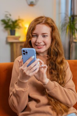 Evdeki oturma odasında gülümseyen cep telefonu kullanan kızıl saçlı mutlu bir genç kadın. Akıllı telefon sosyal medya uygulamalarında internetten film izleyen kız mesajları paylaşıyor.