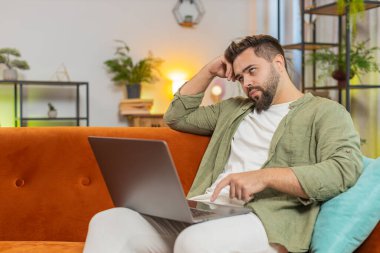 Kahverengi saçlı, aşırı yorgun beyaz adam evdeki kanepede otururken dizüstü bilgisayar kullanmaktan sıkılmış. Uykulu adam işine konsantre olamıyor ve tembelce yazıyor. Yetişkin erkek bir projeyle ilgilenmiyor..