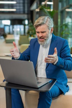 Takım elbiseli kızgın yetişkin patron, ofiste modern işyerinde otururken bilgisayarla video görüşmesi yapıyor. Tatminsiz iş adamı yöneticileri, şirketlerin sorunları hakkında astlarıyla iletişim kuruyor..