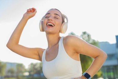 Sporcu spor koşucusu kız antrenman yapıyor kulaklıkla müzik dinliyor, dans ediyor, eğleniyor. Parkta egzersiz kardiyolojisi. Danstan hoşlanan genç bir bayan koşucu. Aktif sporcu kadın