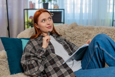 Evdeki genç kadın kanepeye oturup, düşünceli yüzüyle günlüğe yazarak gelecekle ilgili planlarını hayal ediyor. Kafkasyalı kızıl kız rahat, derin duygularla yansıtıyor, hedefleri ve niyetleri belgeliyor.