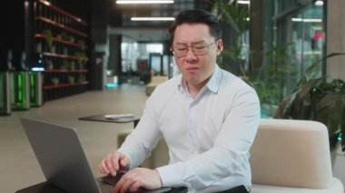 Ofis lobisindeki Çinli işadamı dizüstü bilgisayarla çalışıyor ve diş ağrısının yanağa dokunduğunu hissediyor diş hastalığından dolayı acı veren bir ifade. Pulpitis, sakız hastalığı, diş hastalıkları konsepti.