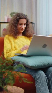 Dizüstü bilgisayar kamerasına bakan çocuk arkadaşlarıyla ya da ailesiyle görüntülü bir konferans görüşmesi yapıyor. 14-15 yaşlarında bir çocuk, evinde, kanepede oturuyor. Dikey