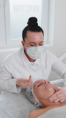 Güzellik uzmanı, genç yetişkin kadın yüzüne nemlendirici tıbbi kimyasallar soyarak krem fırçalama maskesi uyguluyor. Dişi cilt bakımı prosedürü. Yüz kozmetolojisi cilt yenilenmesi. Dikey