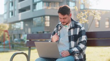 Yetişkin bir iş adamı, şehirdeki bankta otururken bilgisayarla görüntülü konuşma yapıyor. Günlük giysiler içinde erkek yönetici işvereni çevrimiçi konuşma yapıyor, uzaktan iş görüşmesi yapıyor..