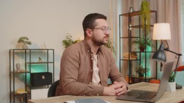 Genç iş adamı dizüstü bilgisayarla oturma odasında kameraya gülümsüyor. Happy Kafkasyalı adam serbest çalışıyor. Ev masasında uzaktan kumandalı çalışıyor. Bilgisayarda E-öğrenme, internette gezinme.