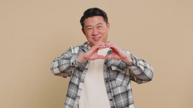 Aşık bir kadın. Mutlu gülümseyen orta yaşlı Asyalı adam kalp hareketi yapar. Aşk işareti, olumlu duyguları ve sempatiyi ifade eder. Olgun Çinli adam bej arka planda izole edilmiş..