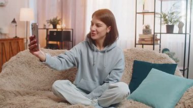 Mutlu genç kadın blogcu akıllı telefondan selfie çekiyor, abonelerle çevrimiçi video görüşmesi yapıyor, sosyal medya için hikayeler kaydediyor. Evde oturan yetişkin kız kanepede oturuyor..