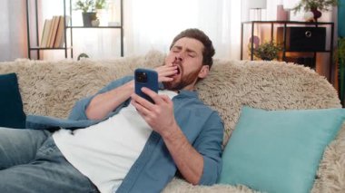 Evdeki genç adam akıllı telefonu elinde tutarken kanepede uyuyakaldı, yorgun hissediyordu. Kafkasyalı adam telefonla dinleniyor, uzun süre ekranda kaldıktan sonra uyanık kalmaya çalışıyor.