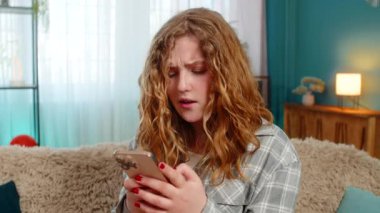 Evde sinirli bir şekilde cep telefonundan mesaj atan genç bir kadın, kızgınlıkla ve hayal kırıklığıyla mesaj yolluyor. Kanepede oturan beyaz saçlı kız el sallıyor, tatminsizliğin duygusal ifadesi.
