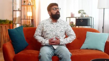 Hintli bir adam acı dolu bir ifadeyle evde oturuyor, midesini tutuyor ve yemekten sonra mide iltihabı ya da mide iltihabı geçiriyor. Arap adam, kanepede dinlenirken karın ağrısı çekiyor.