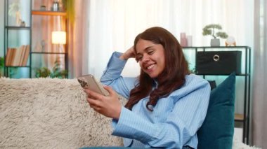 Kanepede oturan Hintli kadın akıllı telefondan komik videolar izliyor ve neşe içinde kahkahalara boğuluyor. Arap kızı evde pozitif duygular ve mutlulukla baş başa zaman geçirmeyi seviyor..