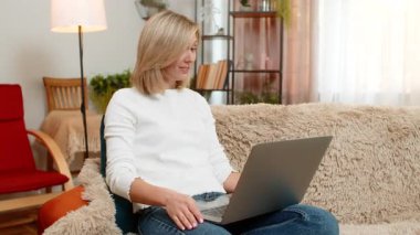 Orta yaşlı bir kadın video toplantısı sırasında evdeki kanepede konuşuyor, gülümsüyor ve anlamlı merhaba jestleri yapıyor. Yetişkin kadın, pozitif enerji ve çatışma göstergesi olarak çevrimiçi ekiple güven içinde iletişim kuruyor..