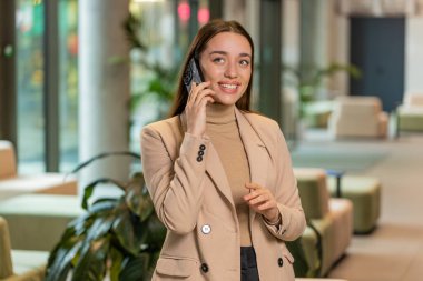 Resmi görevde gülümseyen genç bir iş kadını, modern ofis lobisinde cep telefonuyla konuşuyor. Ulak sohbet uygulamaları kullanarak akıllı telefonu tutan profesyonel yönetici kadın. İstihdam, meslek.