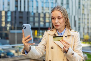 Kafkas 'lı yetişkin iş kadını kredi kartı ve akıllı telefon kullanarak internet üzerinden alışveriş siparişi veriyor. Kız şehir merkezindeki binaya karşı..