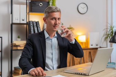 Kafkasyalı bir iş adamı, ofis laptopuyla çalışırken çalışma masasında oturup su içiyor. Takım elbiseli genç serbest çalışan bilgisayar ekranlı sörf internetine bakıyor