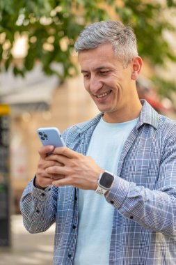 Mutlu, gri saçlı, akıllı telefon kullanan internetteki sosyal medya uygulamalarına girip mesaj yazan açık havada sohbet eden bir adam. Şehir caddesinde duran olgun bir turist. Dikey.