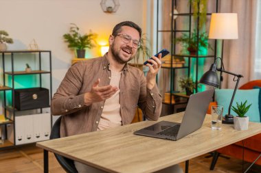 Kafkasyalı genç iş adamı çalışıyor, cep telefonu hoparlörleri ofiste dizüstü bilgisayarla konuşuyor. Kurye sohbet uygulamaları kullanarak akıllı telefonu tutan serbest çalışan bir yönetici. İstihdam, meslek.