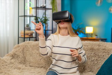 Beyaz kadın evde kanepenin üzerinde sanal gerçeklik oyunu oynarken VR kulaklığı takıyor. Kız heyecanlı, şaşırmış, aktif, kolları hızlı hareket eden, dijital ortama dalmış, şaşkın ve neşeli görünüyor.