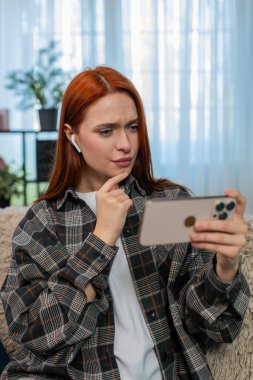 Evde kulaklık takmış akıllı telefondan video izleyen genç bir kadın. Kanepedeki beyaz saçlı kız eğlenceli sosyal medya içeriğinin keyfini çıkarıyor, neşe ve neşeyle tepki veriyor.