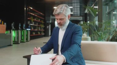 Beyaz tenli orta yaşlı işadamı belgelerle çalışıyor, bilgileri analiz ediyor, gazete okuyor mali rapor hazırlıyor. Takım elbiseli mutlu girişimci ofis lobisindeki grafik belgelerini inceliyor..