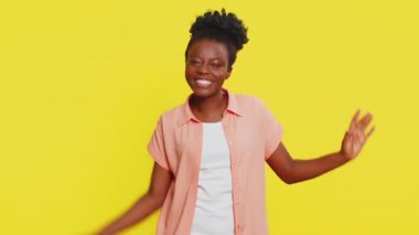 Afrika kökenli Amerikalı bir kadın komik el hareketleriyle dans ediyor, neşe ve neşe saçıyor. Sarı arka planda müzik dinleyen, neşeli ritim ve kaygısız tavır sergileyen siyah bir kız.